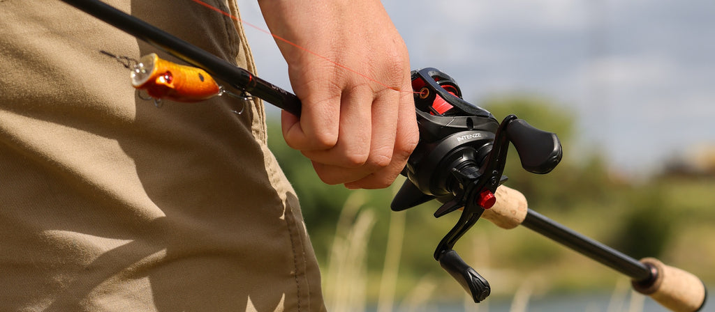 A close-up of a person's hand holding a black DAM QUICK INTENZE baitcasting fishing reel attached to a rod, spooled with red line, with a bright orange and yellow lure visible near the tip. The person is wearing khaki pants, and the background shows a blurred outdoor scene with water and greenery.