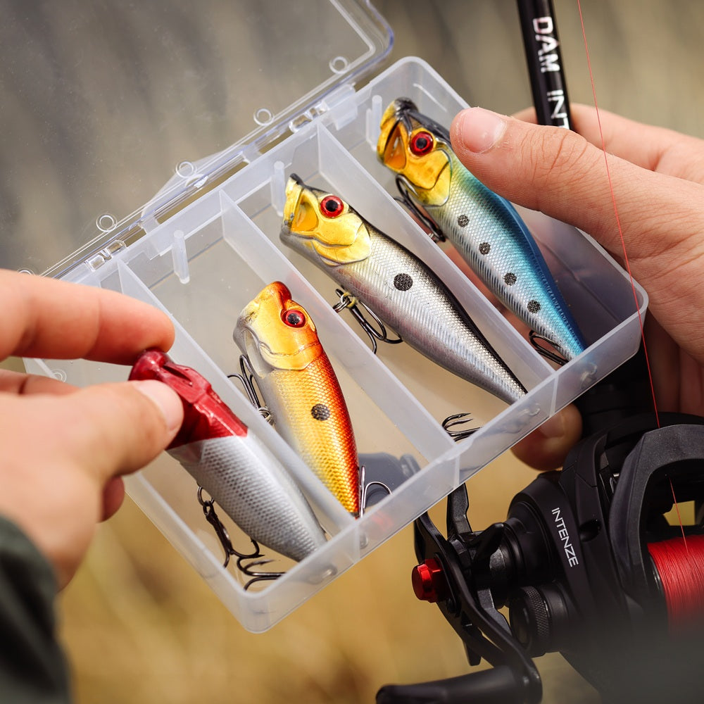 A close-up of a clear plastic fishing lure organizer case holding four DAM hard fishing lures. A person's hands holding the case and a DAM INTENZE fishing rod, with DAM INTENZE Baitcasting Reel, spooled with red line, visible at the right edge.